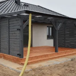 Zadaszony taras z drewnianą podłogą i szarą, drewnianą konstrukcją ścianek bocznych, widoczny wąż doprowadzający wodę i niedokończone obrzeże z kostki brukowej.