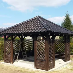 Ciemnobrązowa konstrukcja z drewnianą kratownicą i ciemnym dachem z dachówki, prawdopodobnie w trakcie montażu, z drabiną opartą o jedną ze ścian i widocznym kompresorem obok, na tle błękitnego nieba.