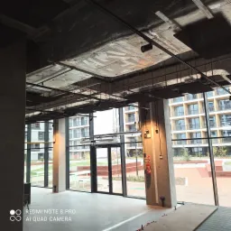 Wnętrze biurowe w trakcie wykańczania, widoczne instalacje pod sufitem, oświetlenie punktowe, system wentylacji, duże okna z widokiem na zewnątrz, betonowe kolumny z przewodami elektrycznymi...