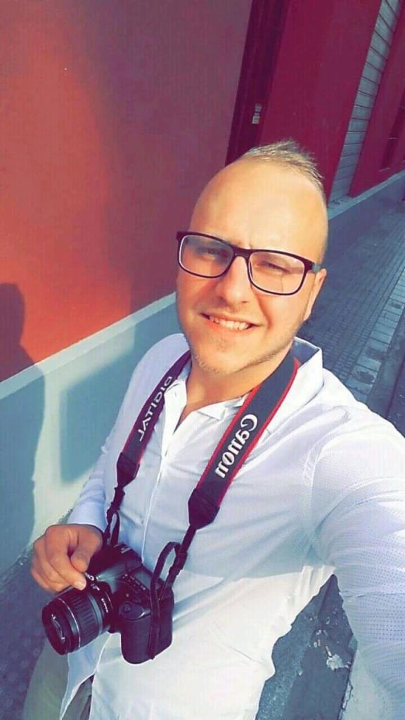 Uśmiechnięty mężczyzna w okularach z aparatem Canon na szyi, ubrany w białą koszulę, pozuje na tle czerwonej ściany i chodnika. Lekkie, boczne oświetlenie.