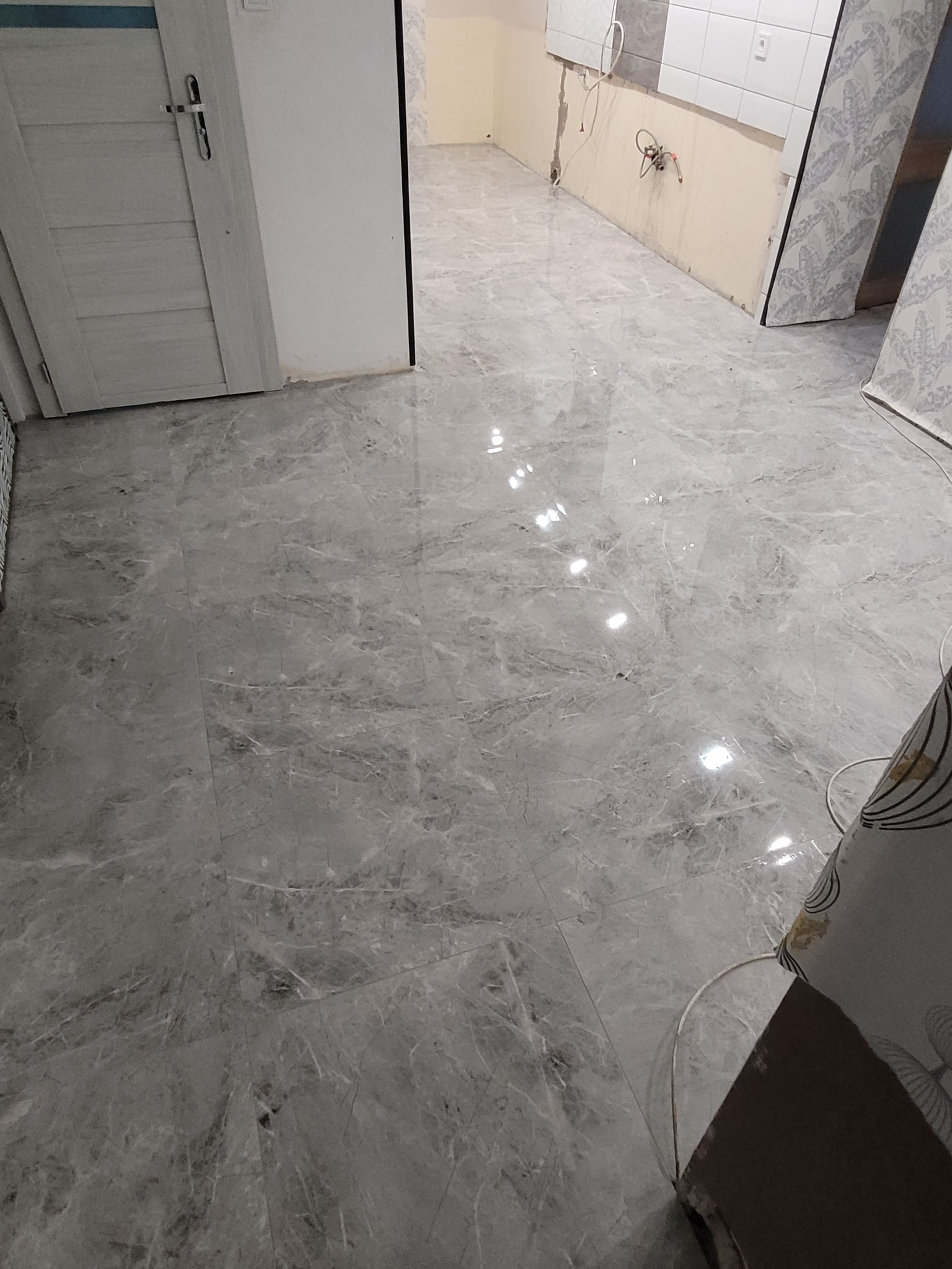 Świeżo położona, błyszcząca, szara marmurowa podłoga z widocznymi refleksami światła, fragmentem białych drzwi po lewej stronie i przygotowaną ścianą pod płytki w tle.