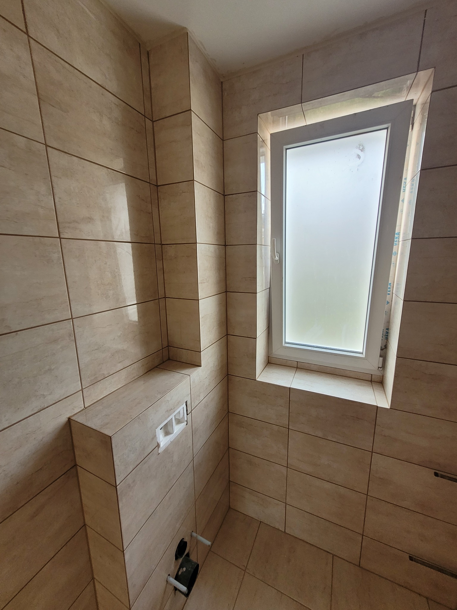 Wykończona łazienka z beżowymi płytkami imitującymi drewno, zabudowany stelaż WC z przyciskiem oraz okno z mleczną szybą.