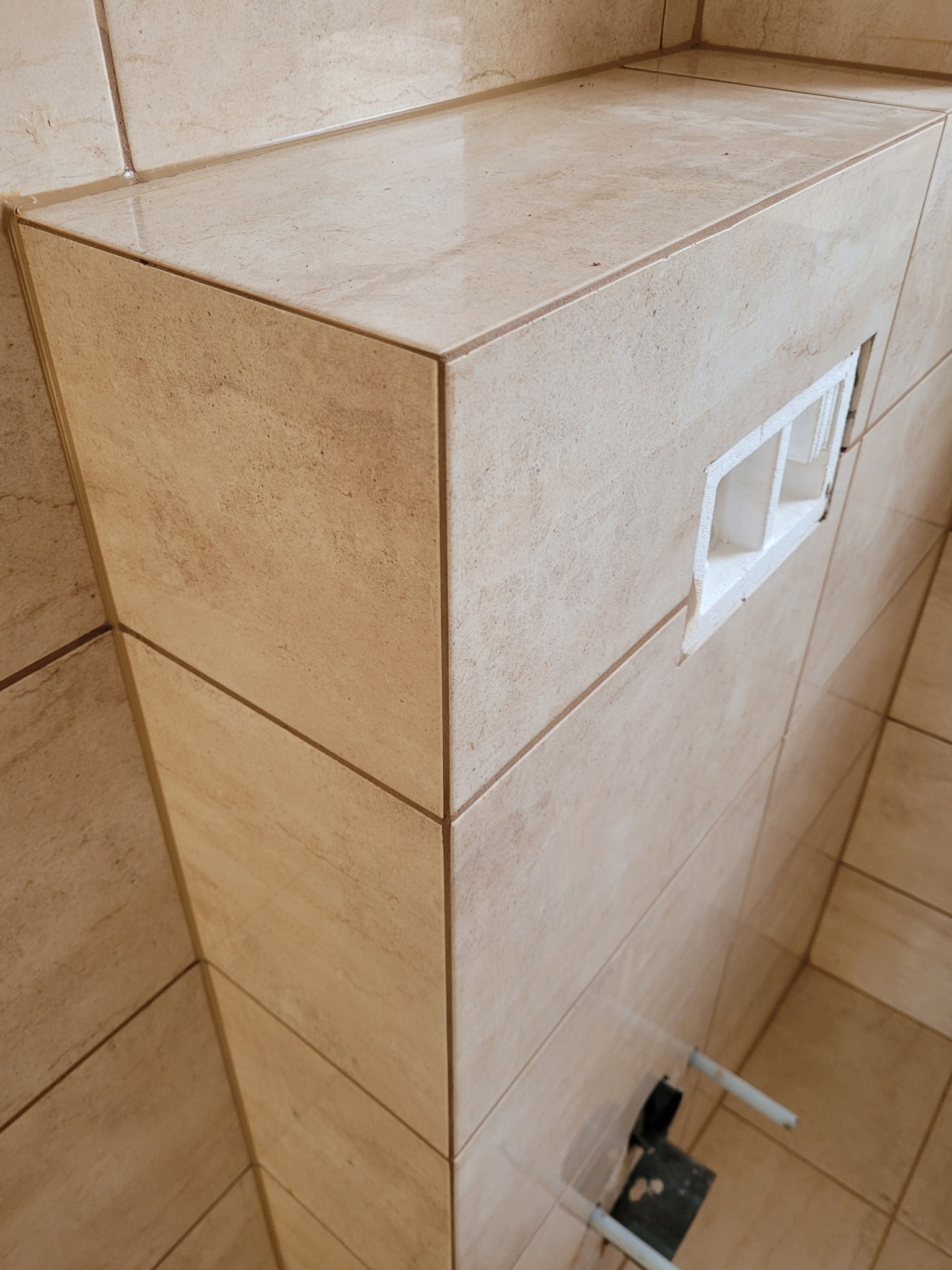 Obudowa stelaża podtynkowego WC wykończona beżowymi płytkami imitującymi kamień z widocznym otworem na przyciski spłukiwania.