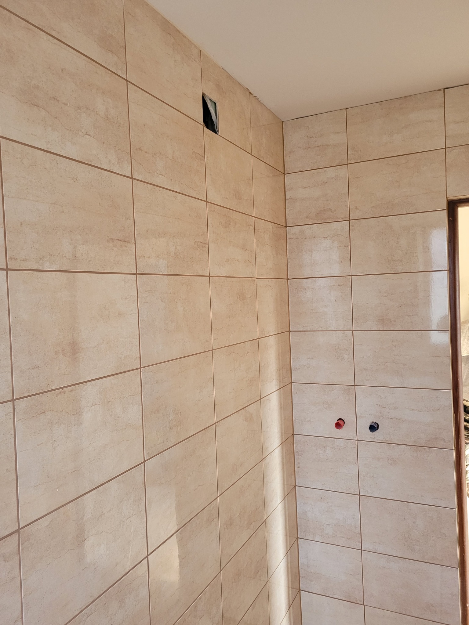 Ściana łazienki w Babimoście wyłożona beżowymi, prostokątnymi płytkami ceramicznymi, z widocznymi otworami na rury i puszką elektryczną, w trakcie prac wykończeniowych.