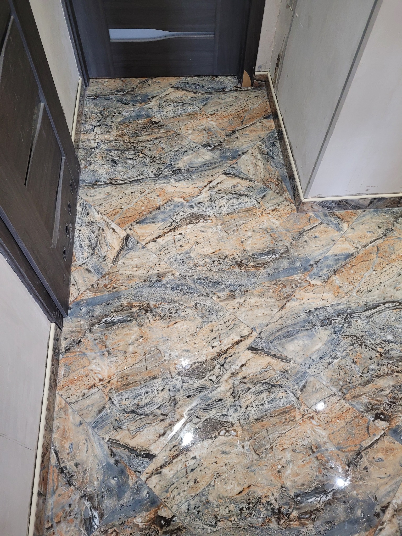 Ułożona podłoga z płytek imitujących marmur w odcieniach beżu, szarości i brązu, widoczne fragmenty ściany i ościeżnicy drzwiowej.