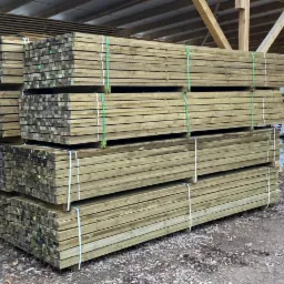 Stosy tarcicy budowlanej ułożone na paletach w magazynie, zabezpieczone zielonymi i białymi taśmami, widoczne płyty OSB w tle.