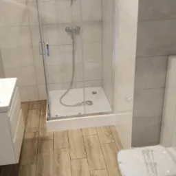 Jasna łazienka po remoncie z kabiną prysznicową, umywalką z szafką, drewnopodobną podłogą i podtynkową spłuczką WC z chromowanym przyciskiem.