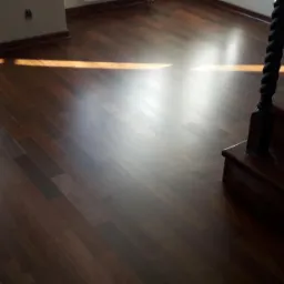 Ciemna, lakierowana podłoga z drewna egzotycznego, ułożona w regularny wzór, z widocznym fragmentem drewnianych schodów z ozdobnymi tralkami i smugą światła słonecznego wpadającą przez okno.