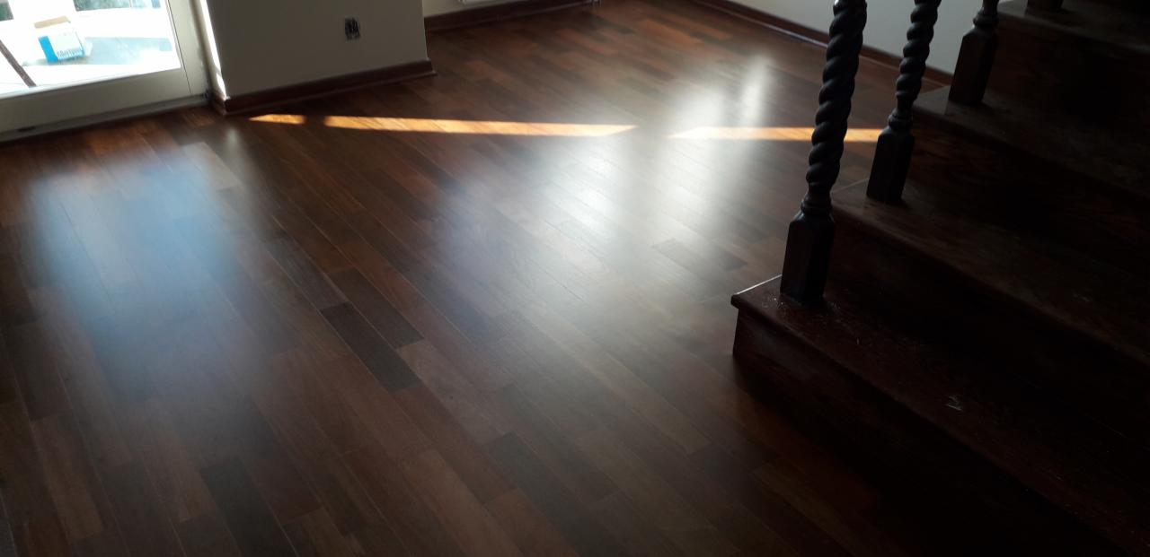 Ciemna, lakierowana podłoga z drewna egzotycznego, ułożona w regularny wzór, z widocznym fragmentem drewnianych schodów z ozdobnymi tralkami i smugą światła słonecznego wpadającą przez okno.