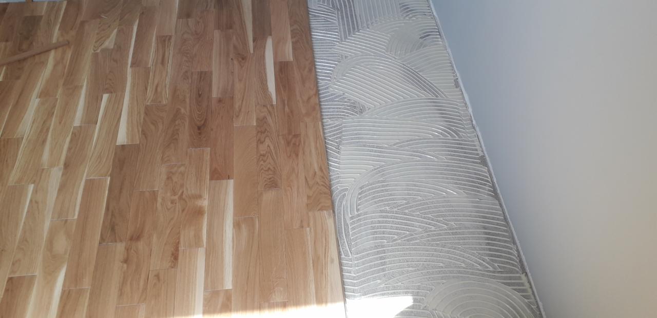 Fragment podłogi: ułożony parkiet z jasnego drewna sąsiaduje z podłożem pokrytym klejem w charakterystyczne fale przed dalszym układaniem.