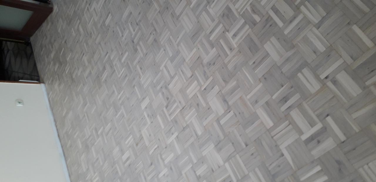 Jasny parkiet ułożony we wzór koszykowy, widoczne usłojenie drewna, fragment ściany i drzwi w tle.