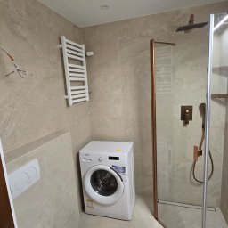 Tomasz Skoczylas - Jasna łazienka z kabiną prysznicową w miedzianej ramie, pralką i grzejnikiem drabinkowym. Beżowe płytki imitujące kamień na ścianach i podłodze.