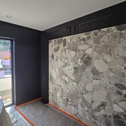 Tomasz Skoczylas - Montaż wielkoformatowych płyt imitujących marmur na ścianie w pokoju z czarnymi ścianami i oknem. Widoczne zabezpieczenia podłogi i okna.