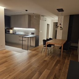 Tomasz Skoczylas - Nowoczesne wnętrze: drewniana podłoga, stół z krzesłami, minimalistyczna kuchnia z oświetleniem LED i dekoracyjną ścianą. Widok na salon z elementami wykończenia.