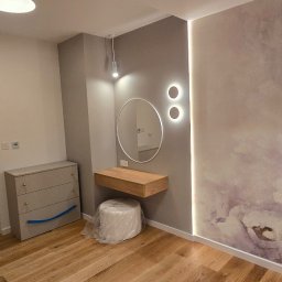 Tomasz Skoczylas - Wykończone wnętrze pokoju z drewnianą podłogą, komodą, toaletką z lustrem i tapetą w kwiaty. Nowoczesne oświetlenie punktowe i listwa LED.