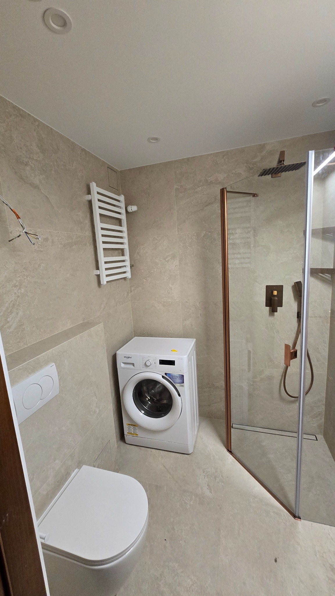 Jasna łazienka z kabiną prysznicową w miedzianej ramie, pralką i grzejnikiem drabinkowym. Beżowe płytki imitujące kamień na ścianach i podłodze.