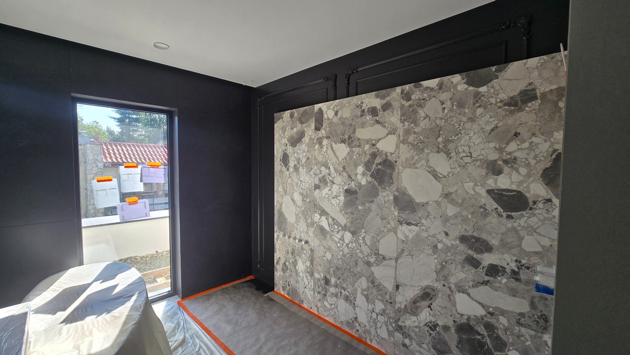 Montaż wielkoformatowych płyt imitujących marmur na ścianie w pokoju z czarnymi ścianami i oknem. Widoczne zabezpieczenia podłogi i okna.