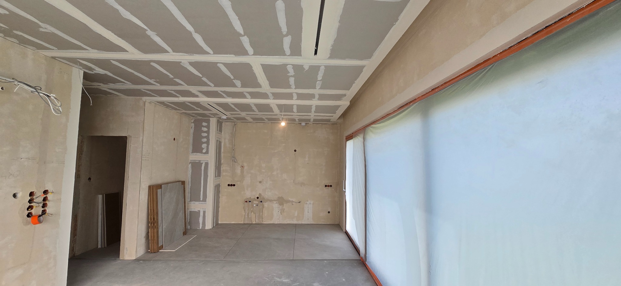 Wnętrze w trakcie prac wykończeniowych: sufit z płyt karton-gips, ściany przygotowane do malowania, widoczne instalacje elektryczne i sanitarne. Okno zabezpieczone folią.