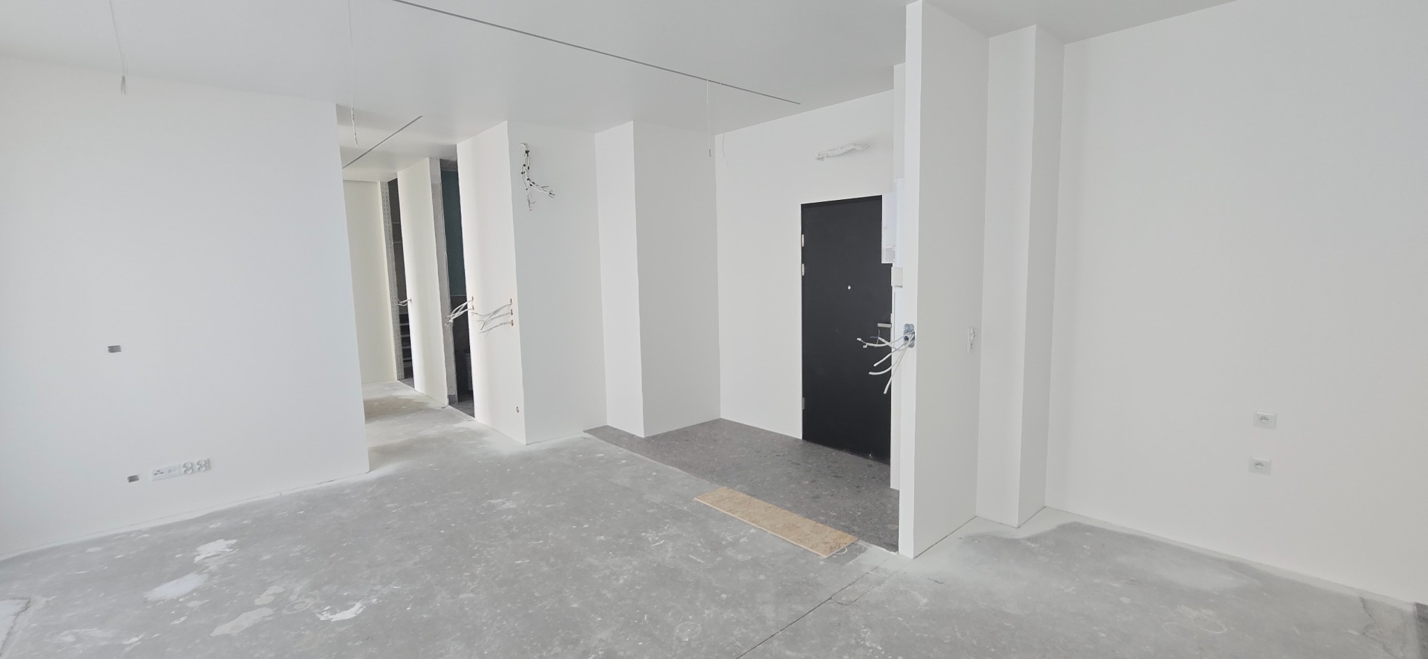 Wnętrze w trakcie wykończenia: białe ściany, surowy beton na podłodze, widoczne kable elektryczne, czarne drzwi wejściowe. Minimalistyczny styl, etap prac wykończeniowych.
