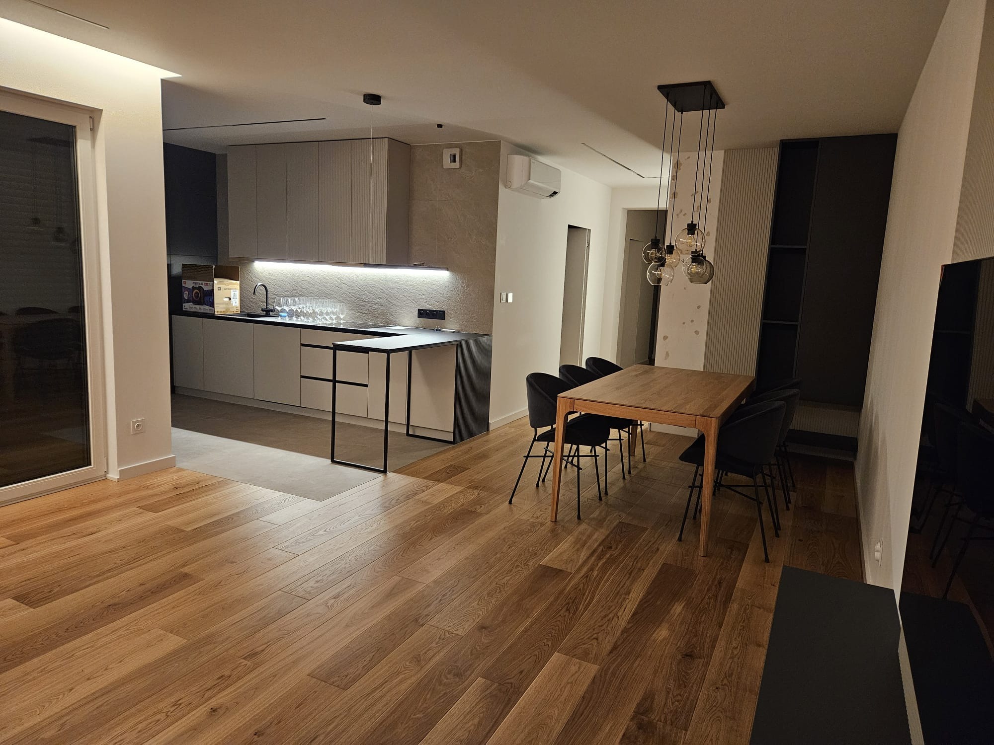 Nowoczesne wnętrze: drewniana podłoga, stół z krzesłami, minimalistyczna kuchnia z oświetleniem LED i dekoracyjną ścianą. Widok na salon z elementami wykończenia.