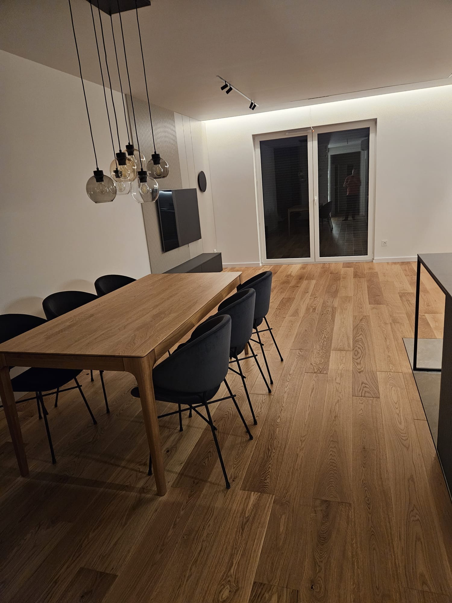 Nowoczesna jadalnia z drewnianym stołem, krzesłami na metalowych nogach i parkietem. Nad stołem designerska lampa z kilkoma szklanymi kloszami. Minimalistyczny wystrój.