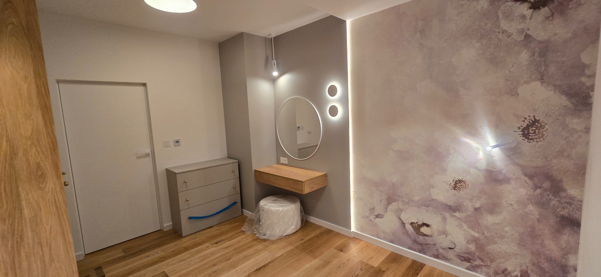 Wykończone wnętrze pokoju z drewnianą podłogą, komodą, toaletką z lustrem i tapetą w kwiaty. Nowoczesne oświetlenie punktowe i listwa LED.