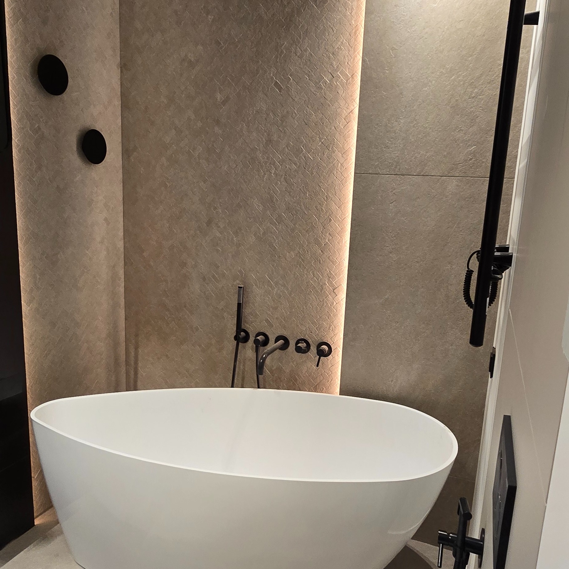 Nowoczesna, wolnostojąca wanna w wykończonej łazience. Beżowe płytki z subtelnym wzorem, czarna armatura i oświetlenie LED tworzą elegancki klimat.