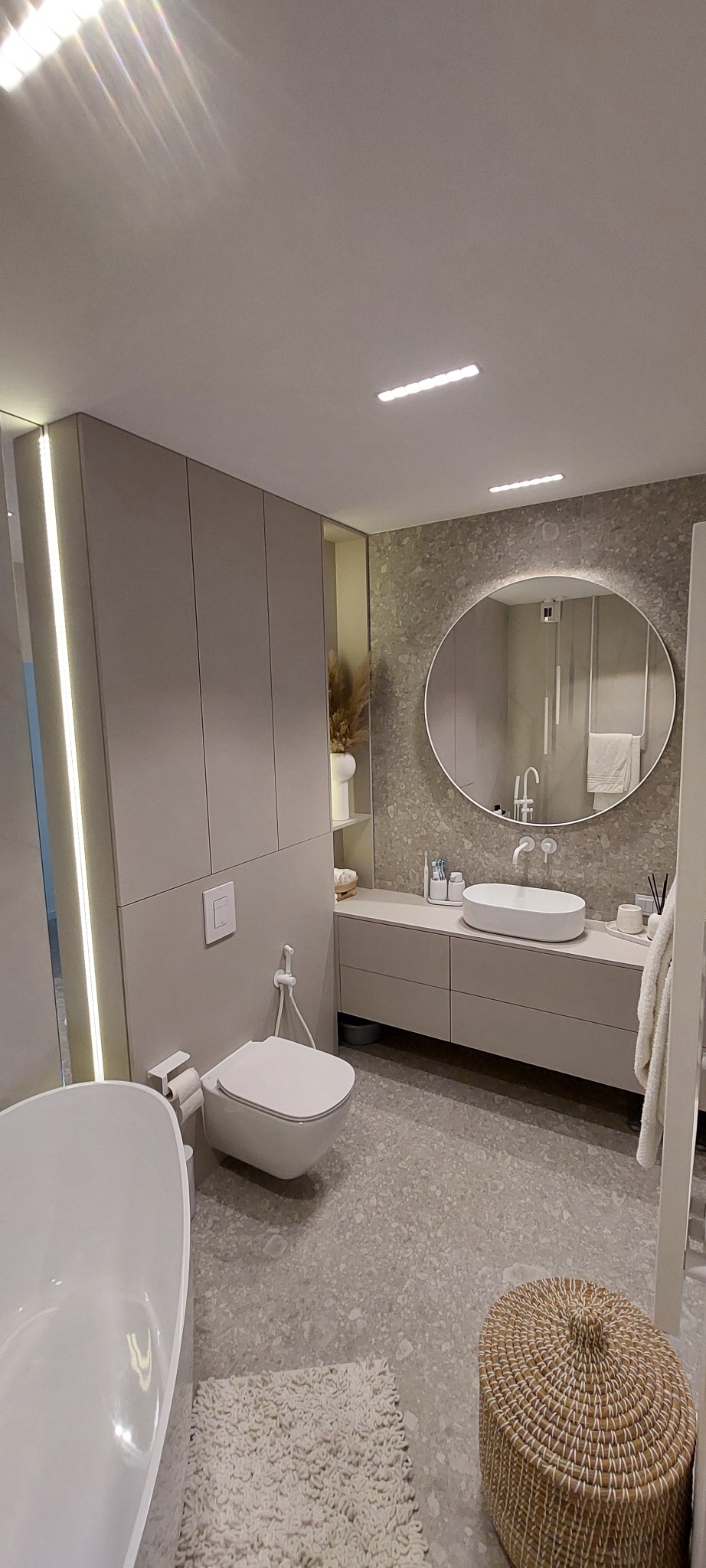 Nowoczesna łazienka w beżach: owalne lustro z podświetleniem, wisząca toaleta, wanna i szafki. Minimalistyczny design z akcentami naturalnych materiałów.