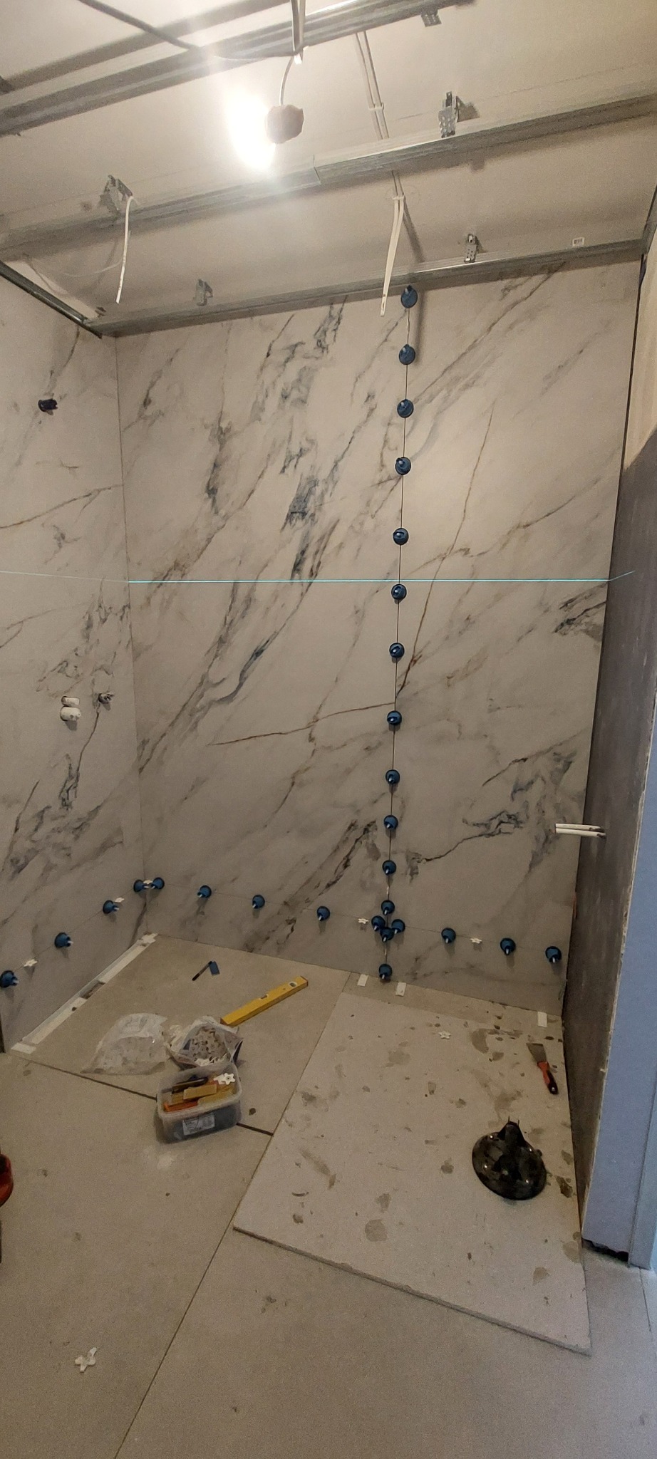 Układanie płytek imitujących marmur w łazience, z użyciem poziomicy i niebieskich krzyżyków dystansowych. Widoczne narzędzia i fragment sufitu podwieszanego.