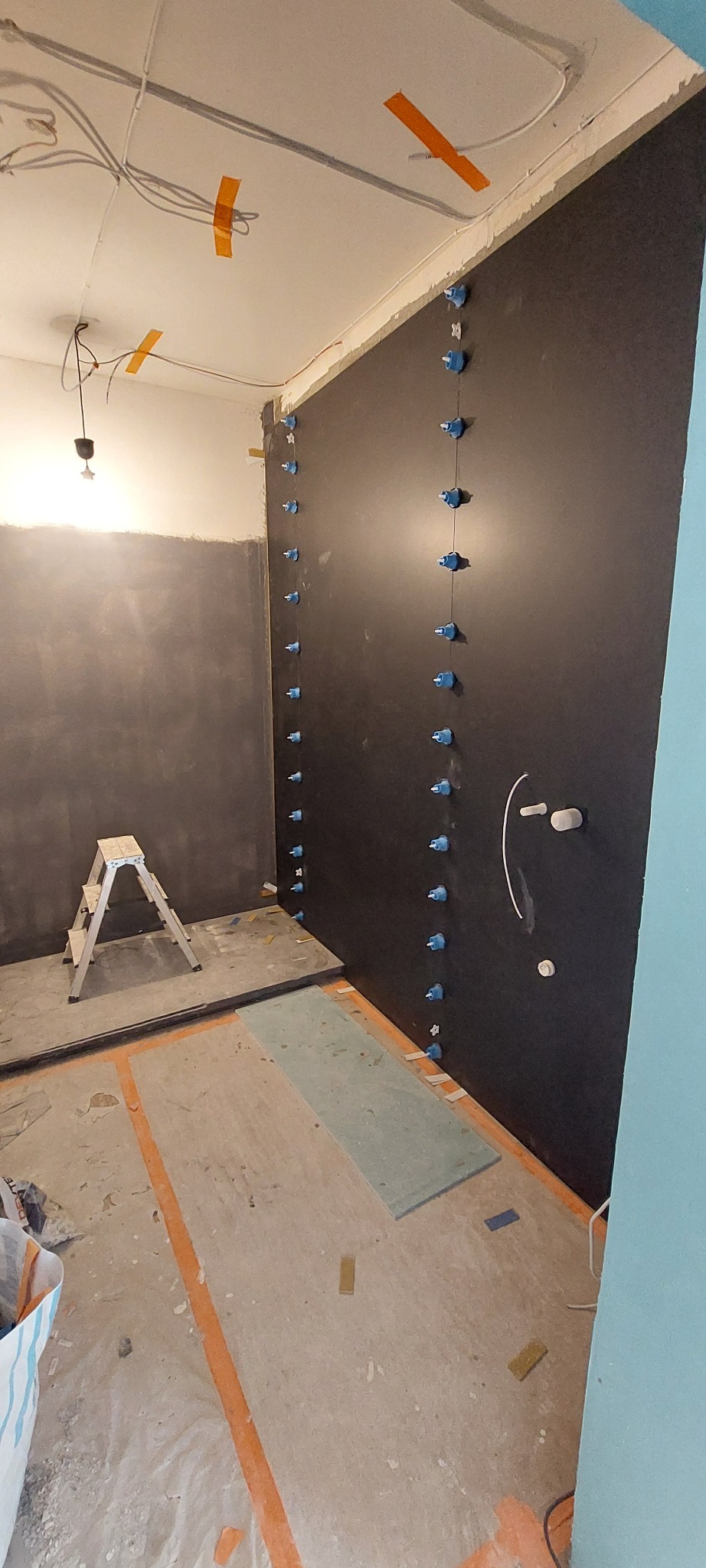 Montaż czarnej płyty karton-gips na ścianie w łazience, z użyciem niebieskich uchwytów poziomujących. Widoczne instalacje i drabina. Przygotowanie do wykończenia.