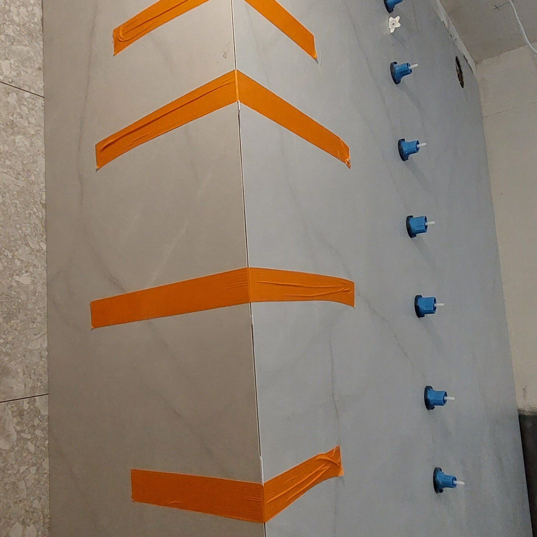 Ściana wyłożona szarymi płytkami, zabezpieczona pomarańczową taśmą i niebieskimi klipsami poziomującymi, ukazująca etap prac glazurniczych w rogu pomieszczenia.