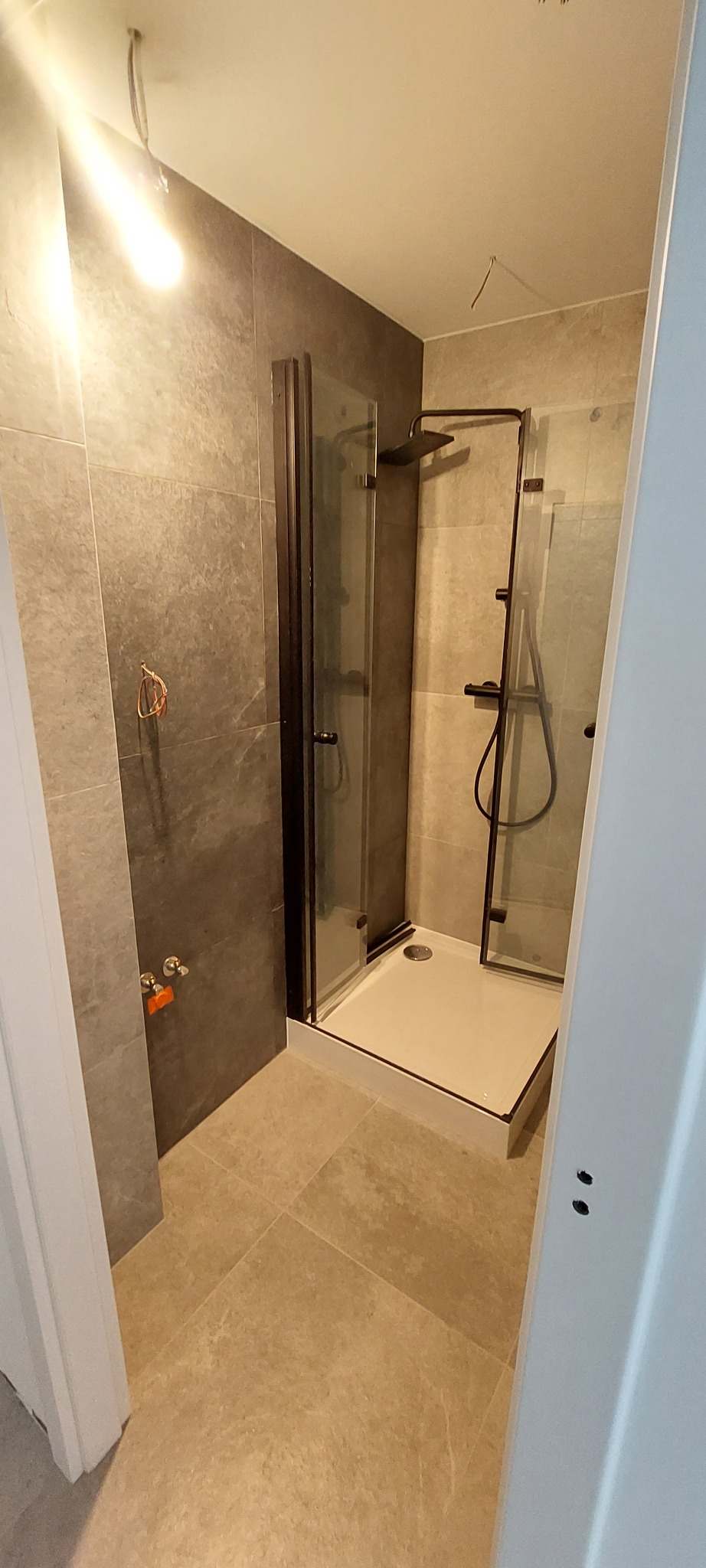 Nowoczesna kabina prysznicowa z czarnymi profilami i deszczownicą, wykończona płytkami w odcieniach szarości i beżu. Widoczne przyłącza wodne i fragment otwartych drzwi.