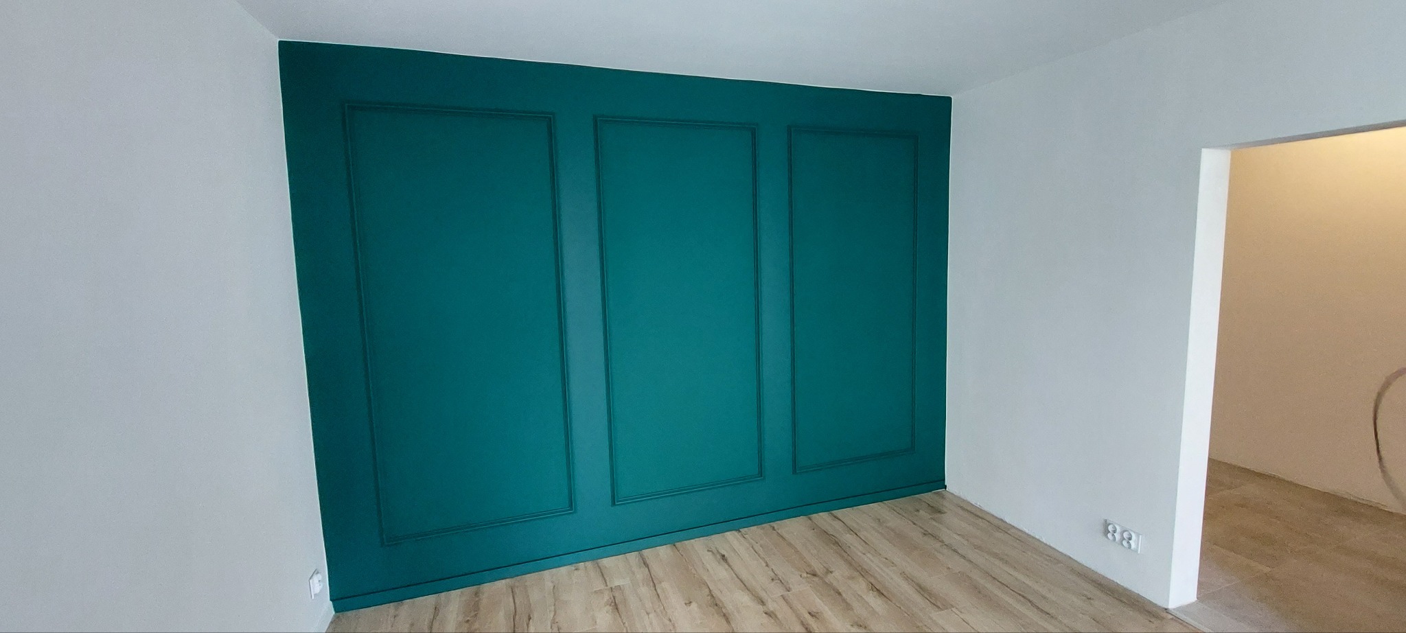 Ściana z dekoracyjnymi panelami w odcieniu szmaragdowej zieleni, kontrastująca z białymi ścianami i jasną podłogą. Widoczny fragment otwartego przejścia do innego pomieszczenia.