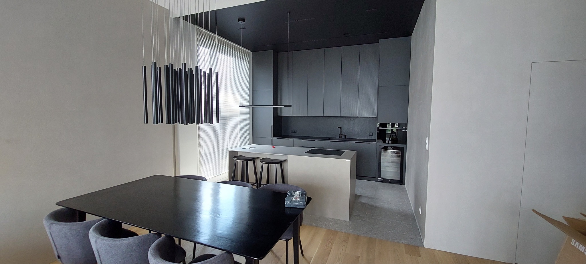 Nowoczesna kuchnia w odcieniach szarości z wyspą i minimalistycznym oświetleniem. Widoczny stół z krzesłami i skrzynka narzędziowa na blacie.