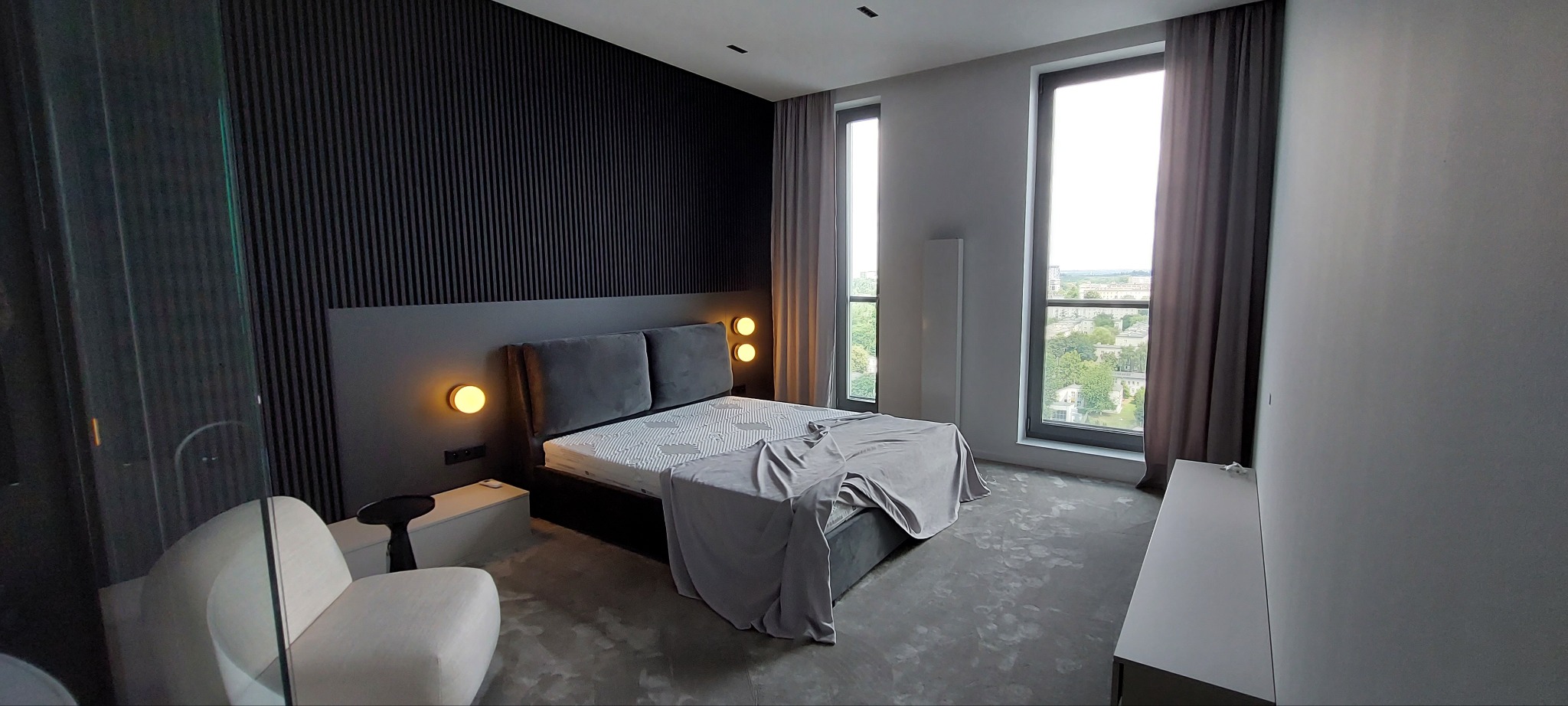 Nowoczesna sypialnia w odcieniach szarości i czerni. Dekoracyjna ściana z pionowych paneli, łóżko z podświetleniem i widok na miasto z okien.