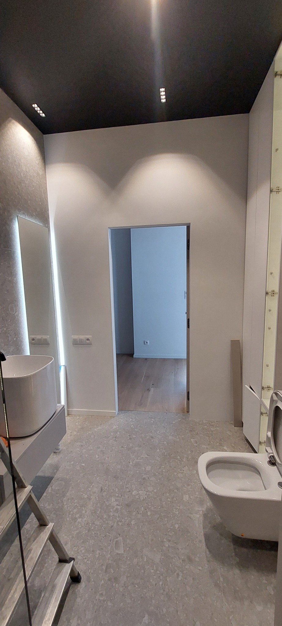 Nowoczesna łazienka w trakcie wykańczania: biała umywalka, toaleta i szara podłoga z terazzo. Widoczna drabina malarska i otwarte drzwi do innego pomieszczenia.