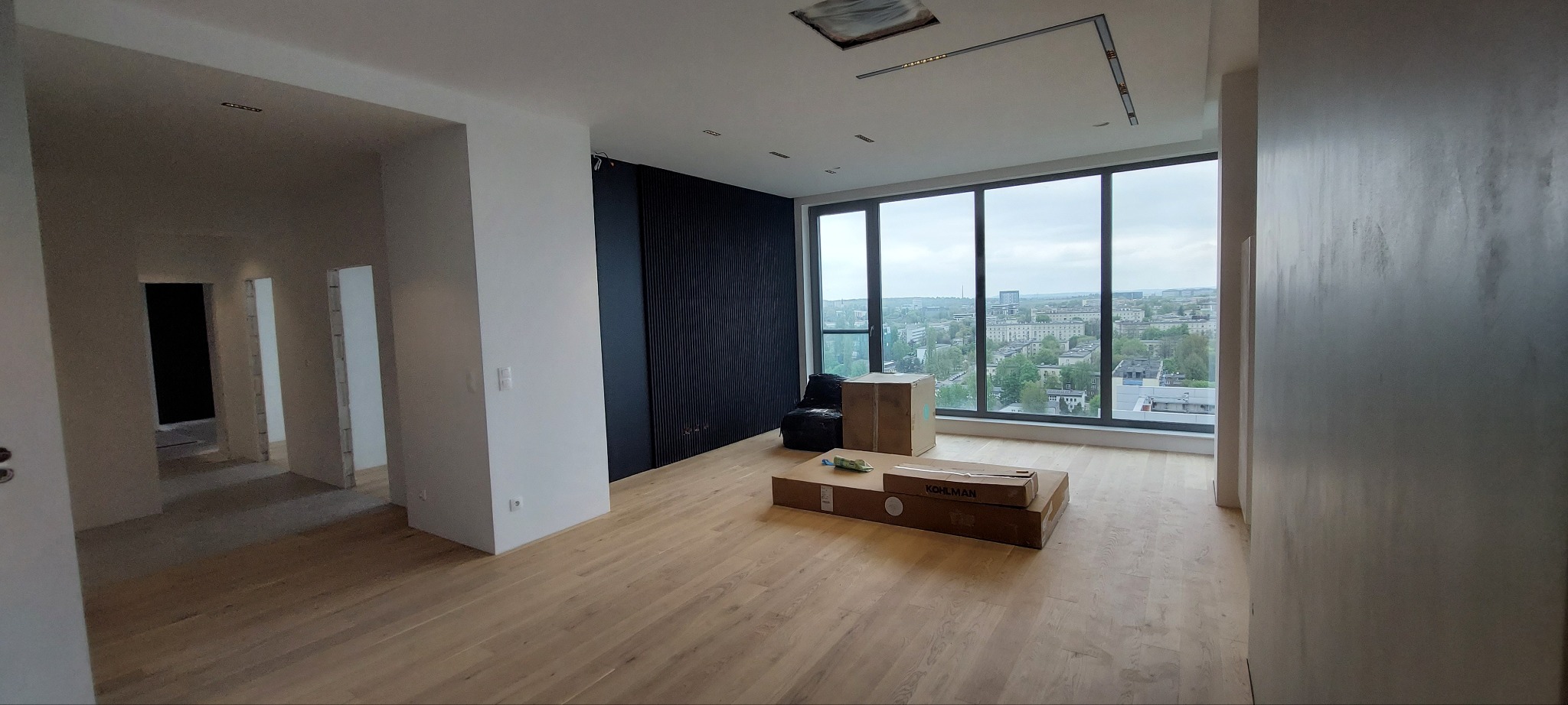 Jasne panele podłogowe w przestronnym wnętrzu z dużymi oknami i widokiem na miasto. Ściany w odcieniach bieli i szarości, czarna ściana dekoracyjna. Kartony na podłodze.