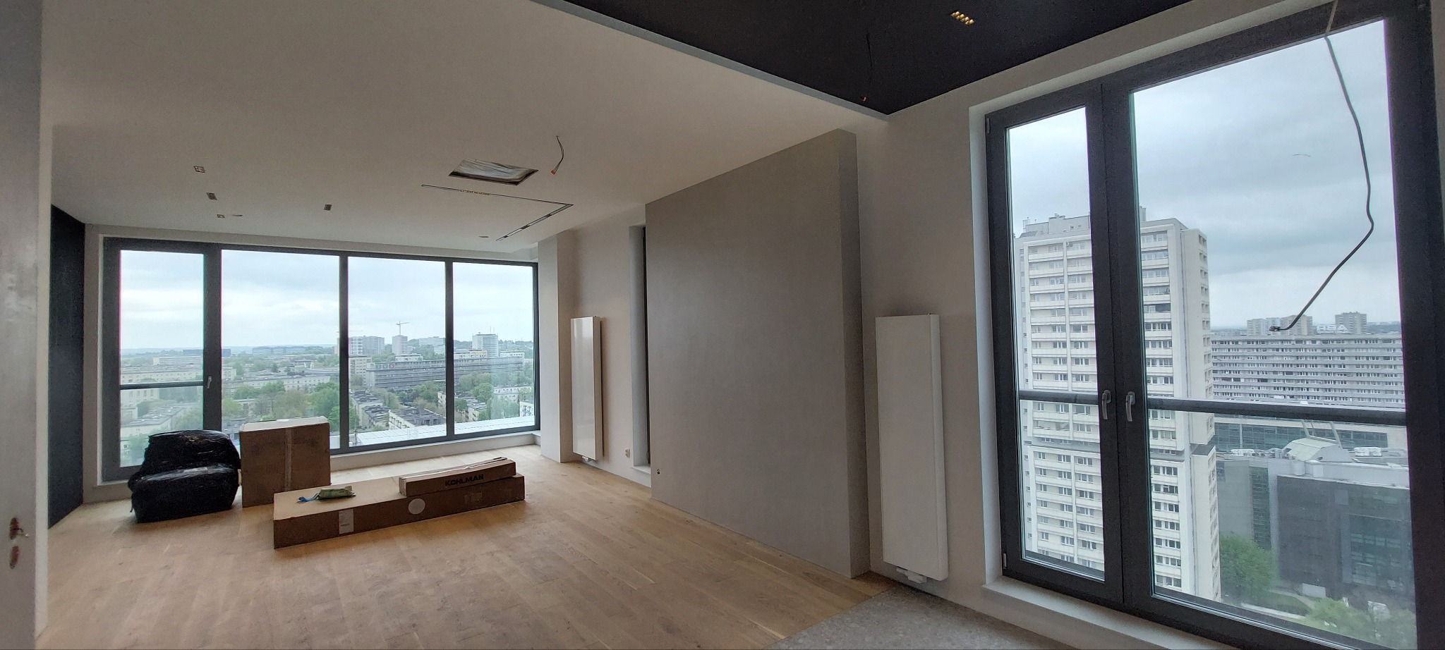 Wnętrze apartamentu w Katowicach w trakcie wykańczania. Widoczne duże okna z widokiem na miasto, podłoga i przygotowane materiały. Oświetlenie sufitowe w trakcie montażu.