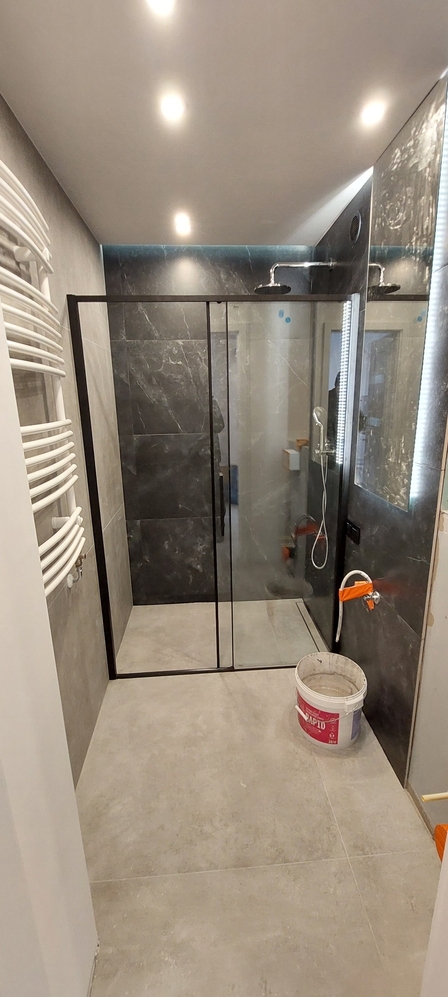 Nowoczesna kabina prysznicowa z czarnymi profilami i deszczownicą, wykończona płytkami imitującymi marmur. Widoczny grzejnik drabinkowy i wiadro z zaprawą.