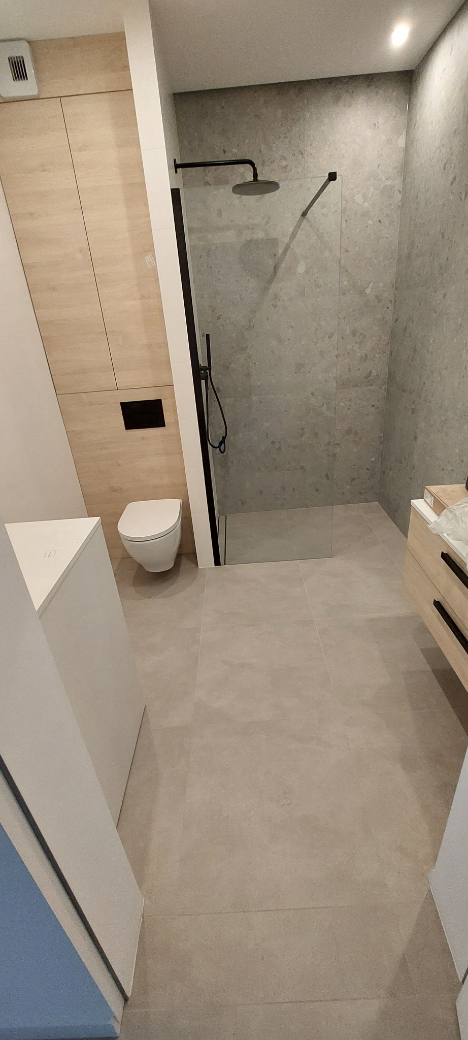 Nowoczesna łazienka z kabiną typu walk-in, szarą mozaiką na ścianach i drewnianymi akcentami. Uwagę zwraca minimalistyczna toaleta podwieszana i czarna armatura.