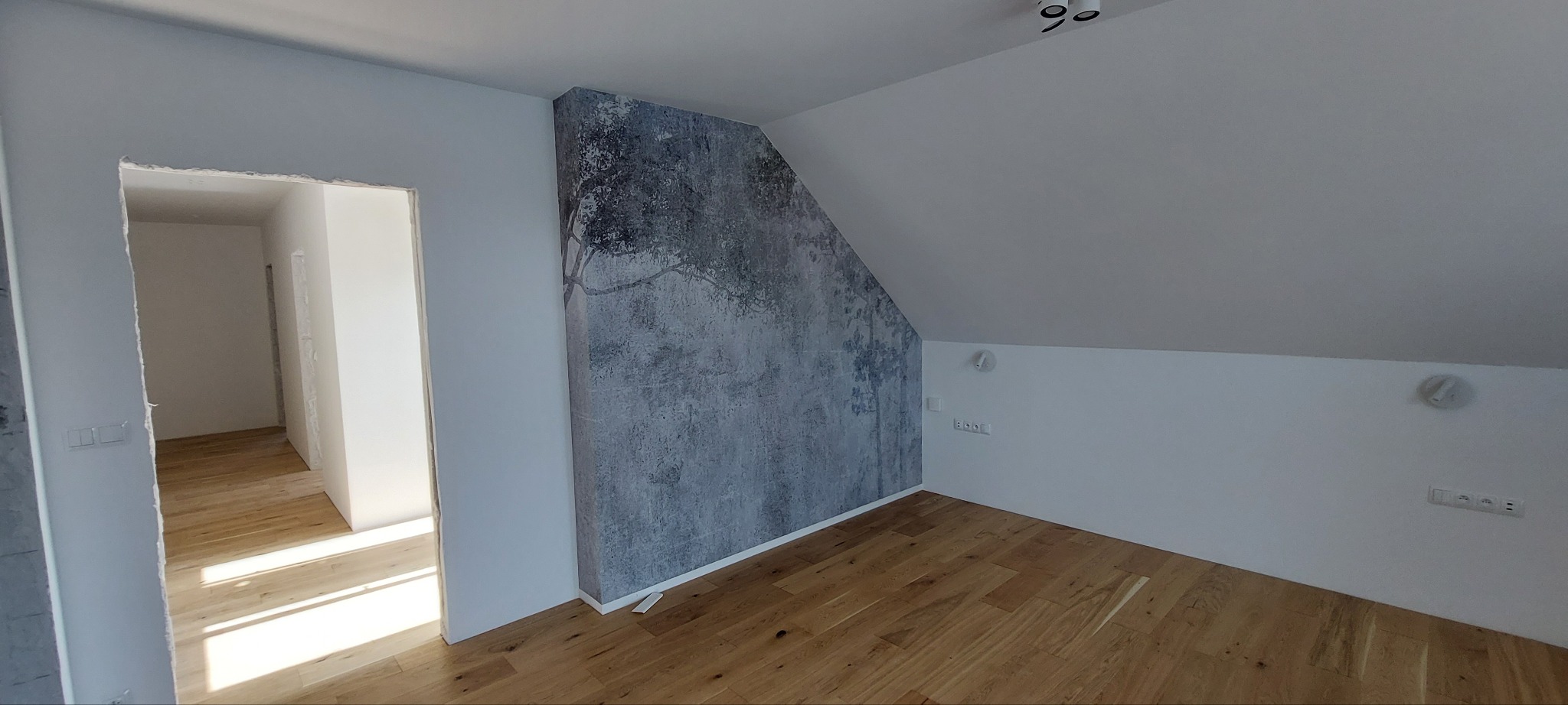 Poddasze po remoncie: drewniana podłoga, ściana z fototapetą z motywem drzewa, widoczne gniazdka elektryczne i oświetlenie sufitowe. Widok na sąsiednie pomieszczenie.