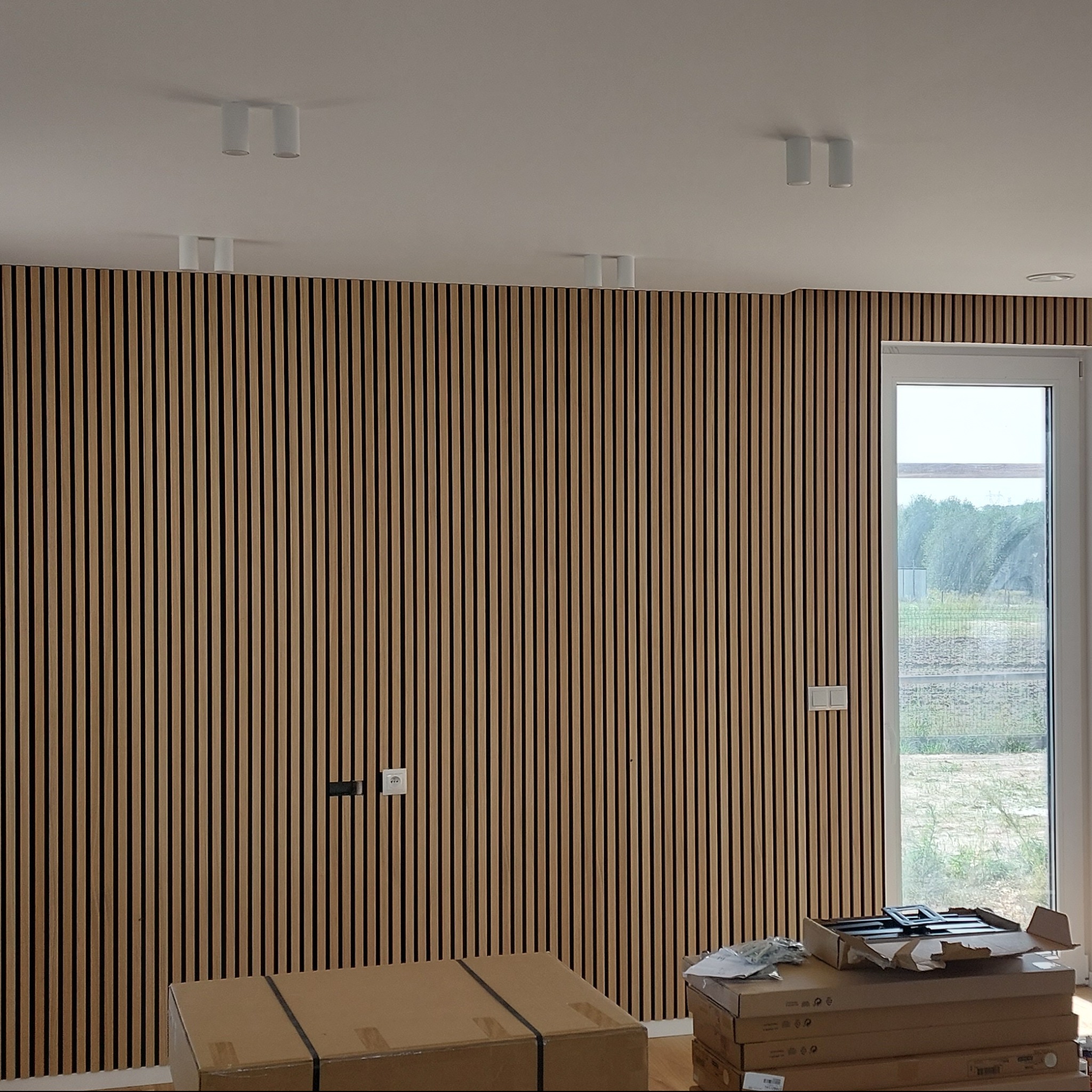 Ściana wykończona panelami lamelowymi w odcieniach beżu i brązu, z widocznymi gniazdkami elektrycznymi i oknem. Na podłodze kartony z elementami wykończeniowymi.