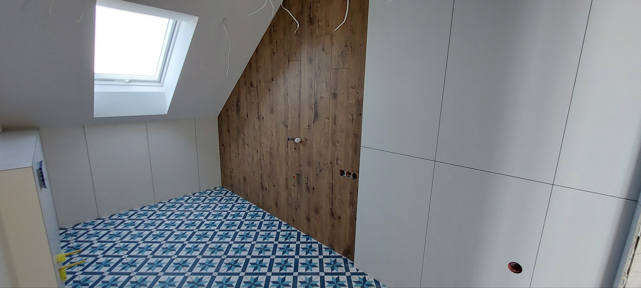Wykończone poddasze ze skosem: drewniana ściana, białe szafy w zabudowie i wzorzysta, niebiesko-biała podłoga. Widoczne gniazdka elektryczne i okno dachowe.