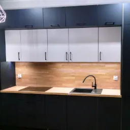 Nowoczesna kuchnia z czarnymi szafkami dolnymi i górnymi, białymi szafkami wiszącymi, drewnianym blatem i panelami ściennymi w odcieniach drewna, oświetlona lampą wiszącą w kształcie diamentu.