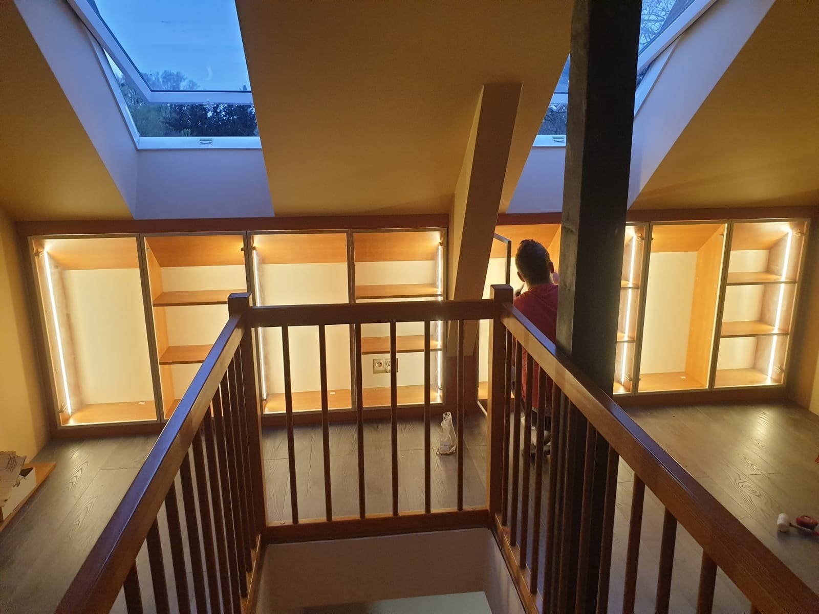 Poddasze z drewnianą balustradą schodów i zabudową szaf z podświetleniem LED, widok z góry na osobę pracującą nad wykończeniem wnętrza szafy.