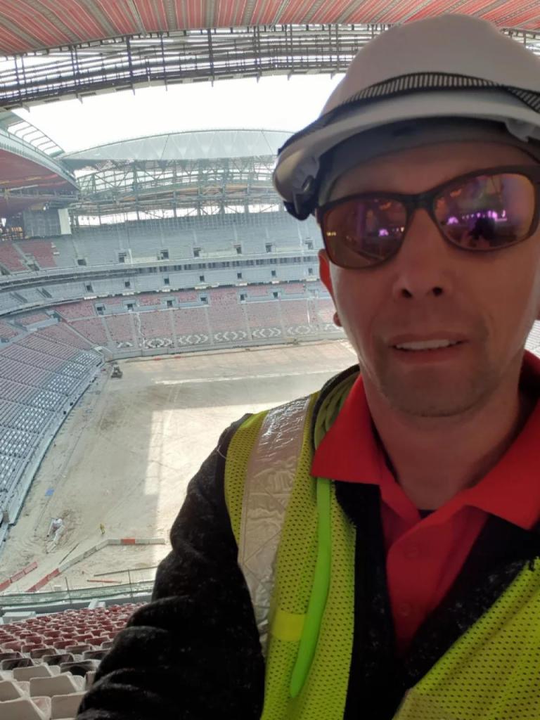 Pracownik w kasku i kamizelce odblaskowej na tle budowy stadionu z widocznymi trybunami i konstrukcją dachu. Ujęcie z góry.