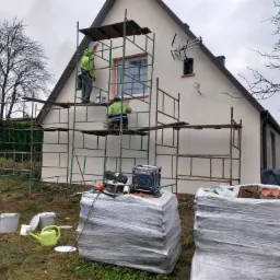 Dwóch pracowników na rusztowaniu maluje elewację domu jednorodzinnego, widoczne narzędzia, pędzle, wiaderka z farbą, oraz zapakowane materiały budowlane.