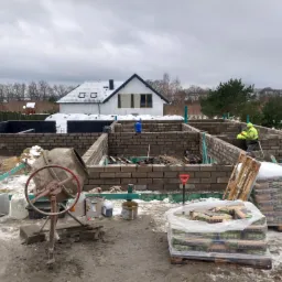 Budowa domu z bloczków: fundamenty, ściany parteru w trakcie murowania, widoczne worki z zaprawą Kreisel, betoniarka i pracownicy w odblaskowych kamizelkach na tle zimowego krajobrazu i częściowo...