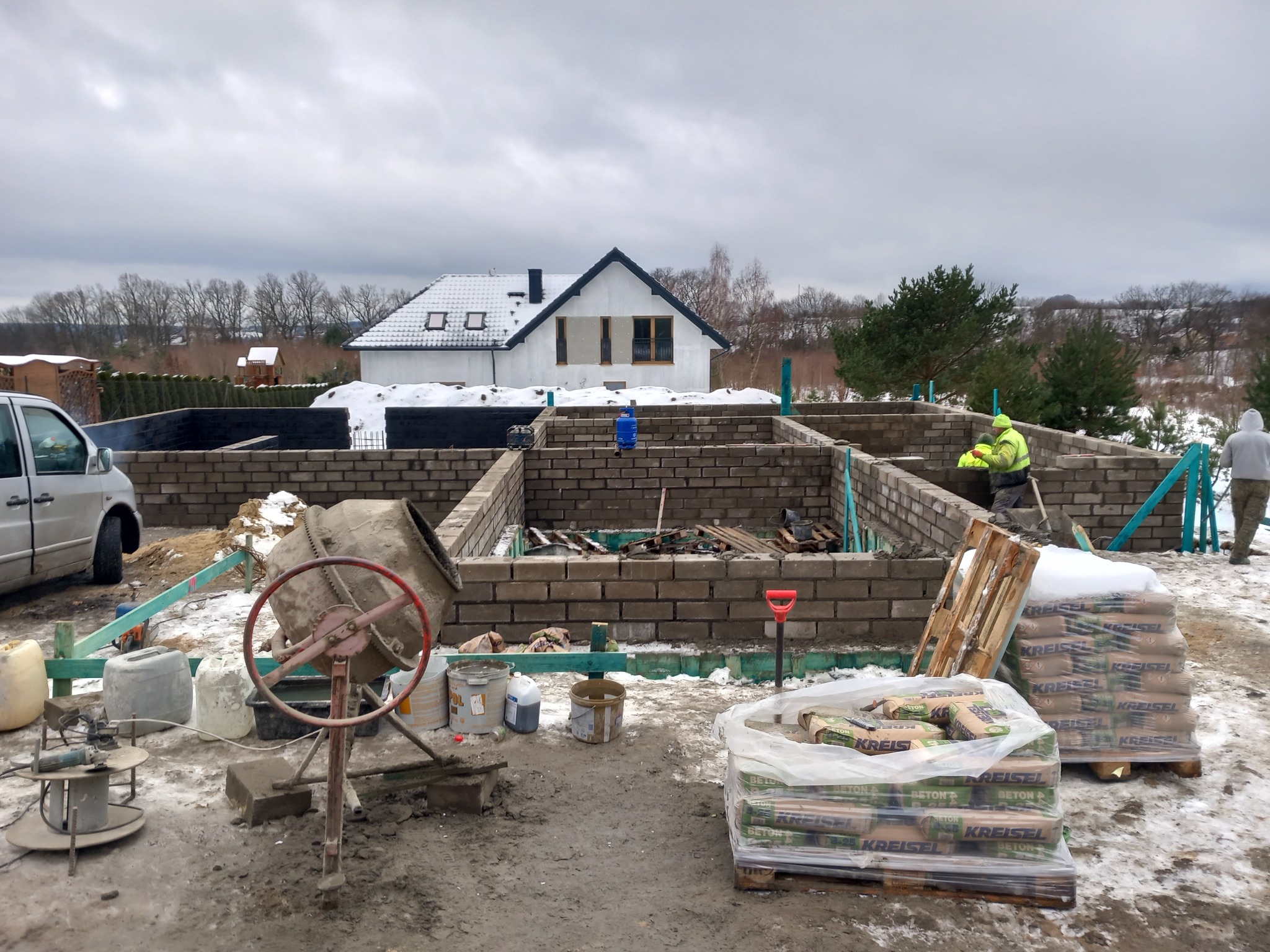 Budowa domu z bloczków: fundamenty, ściany parteru w trakcie murowania, widoczne worki z zaprawą Kreisel, betoniarka i pracownicy w odblaskowych kamizelkach na tle zimowego krajobrazu i częściowo...