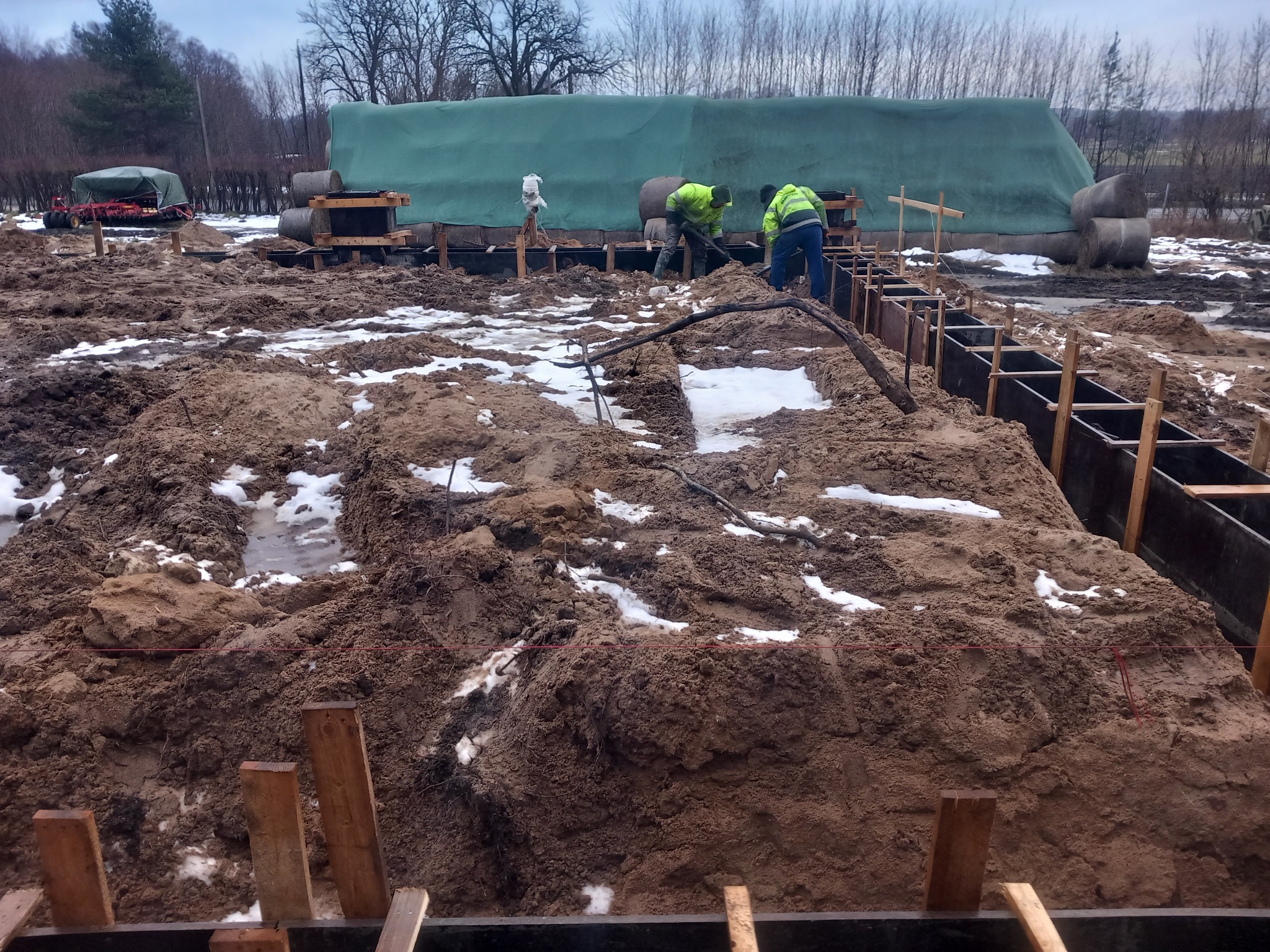 Dwóch robotników w jaskrawozielonych kurtkach pracuje przy wykopie fundamentów pod budynek, widoczne drewniane szalunki i ziemia z resztkami śniegu.